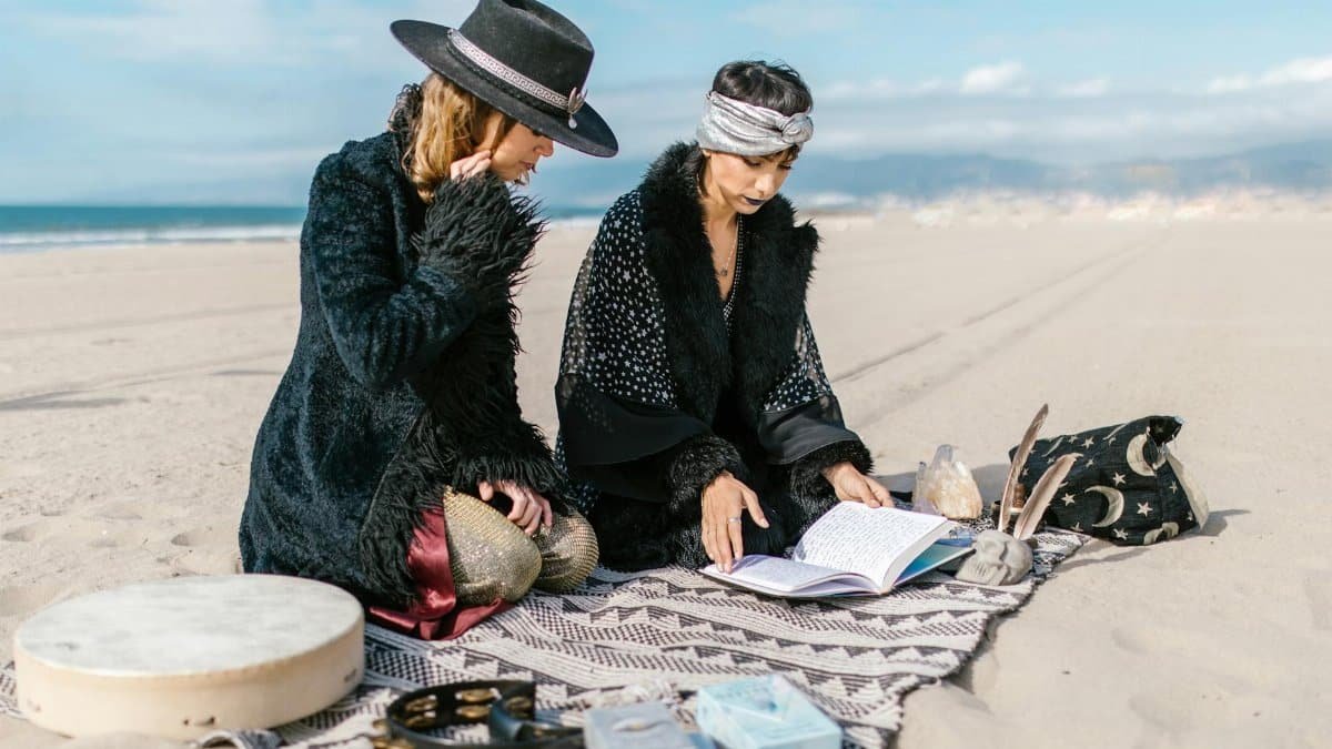 Two women performing a spiritual ritual on a sandy beach with crystal and book.