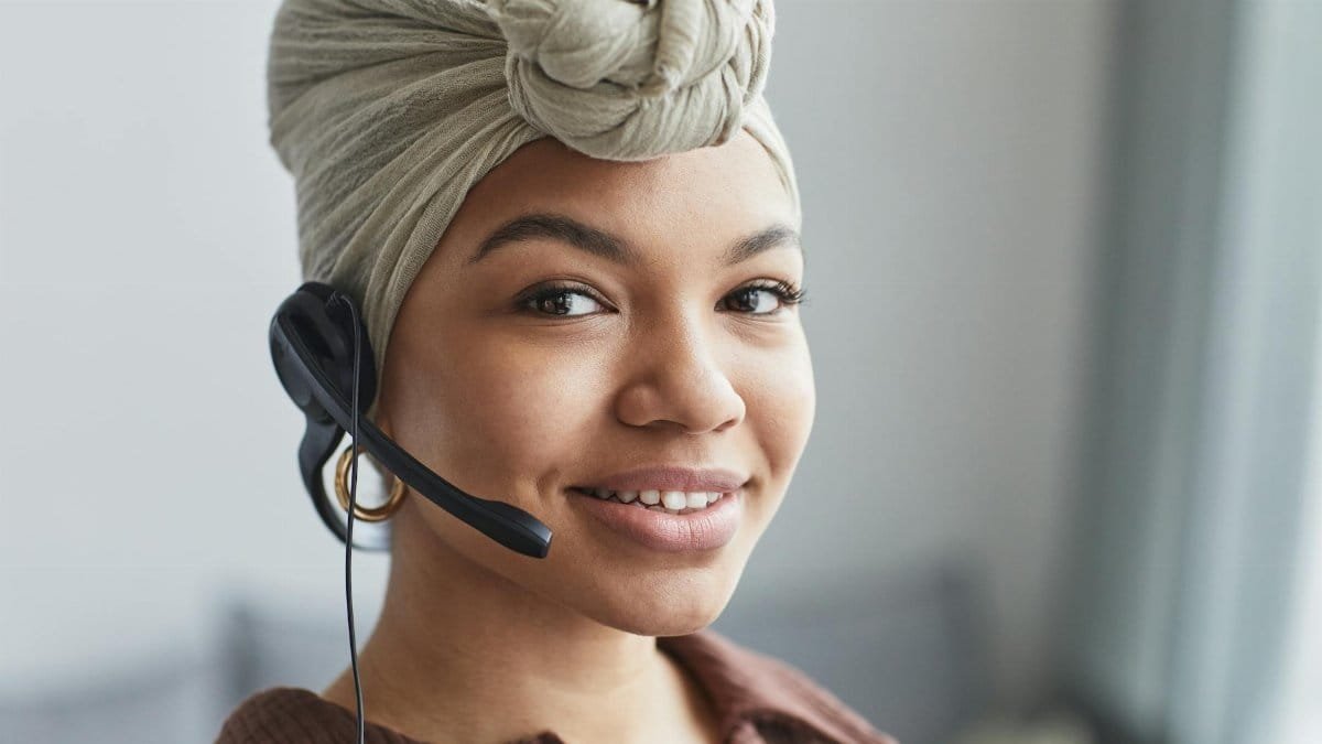 Delighted African American female call agent with headset wearing turban looking away while standing in light room on blurred background