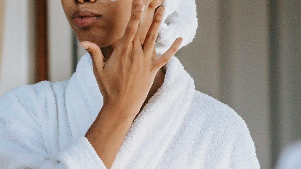 Crop anonymous female in white bathrobe with towel on head applying facial moisturizing cream on face while standing in bathroom