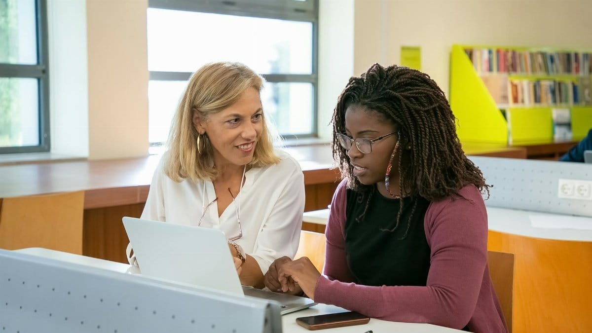 Positive adult female teacher explaining task to young black student with Afro braids doing assignment on laptop in modern classroom
