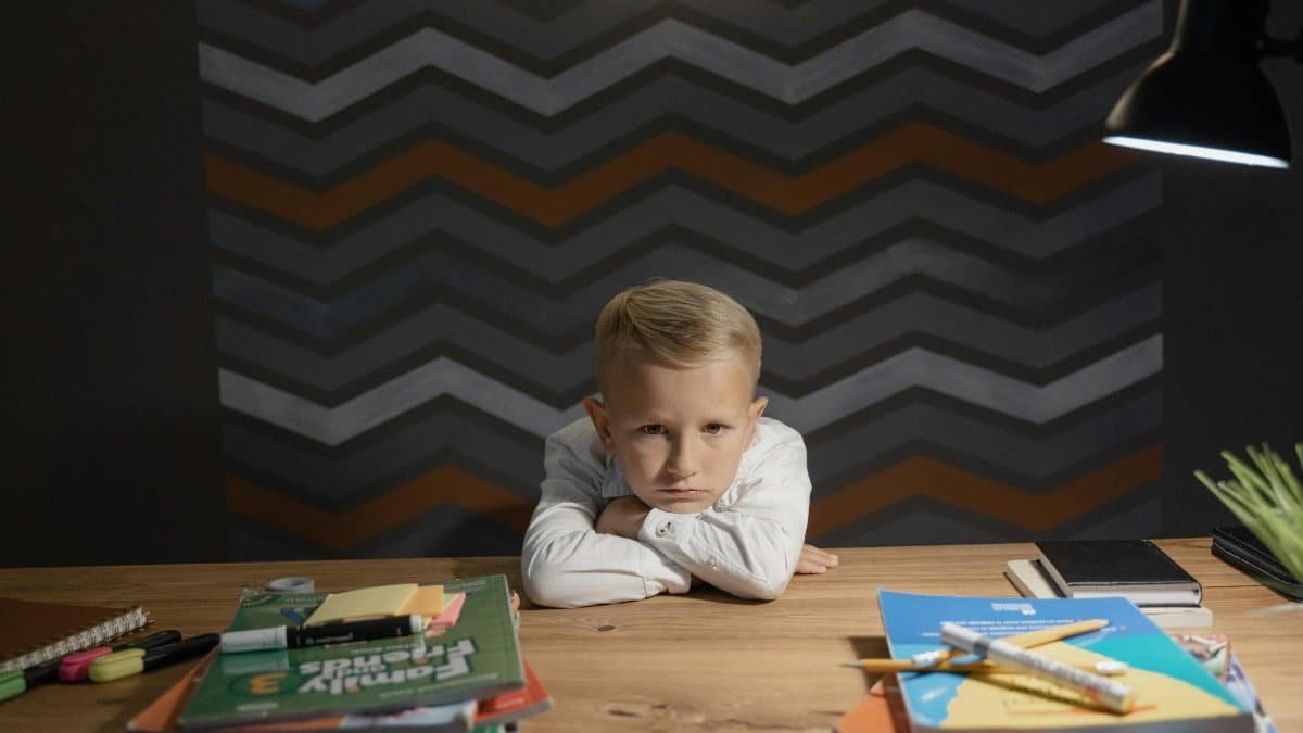A young boy leaning on a table surrounded by books and coloring materials, looking unhappy with studying.
