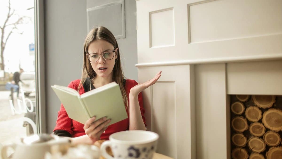 A woman reading and reacting to a book in a cozy cafe, surrounded by coffee and teapots.