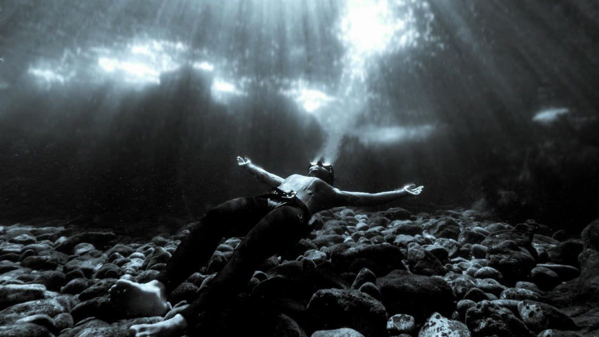 Monochrome photograph of a man floating underwater on a rocky sea bed, illuminated by sunlight.