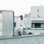 A man in a white suit with angel wings sits on a rooftop, offering a celestial and urban contrast.
