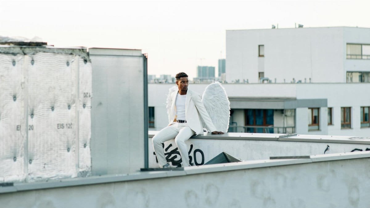 A man in a white suit with angel wings sits on a rooftop, offering a celestial and urban contrast.