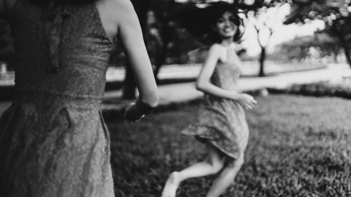 Two women in dresses joyfully run barefoot through a sunlit park meadow.