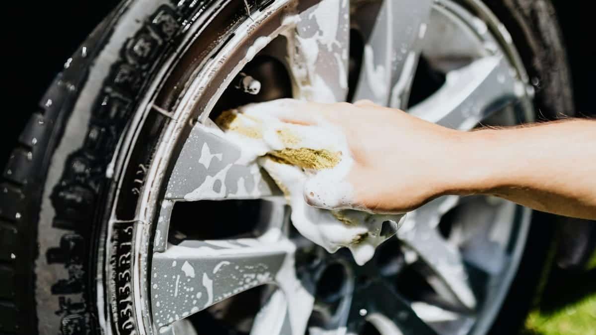 A close-up image of a hand scrubbing a car rim and tire with suds, emphasizing car wash detail.