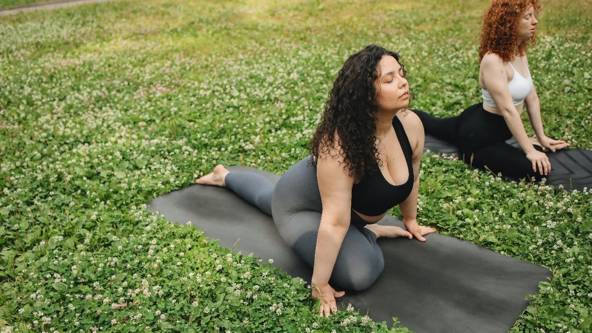 Two women in activewear practicing yoga on mats in a grassy field. Wellness and outdoor fitness.