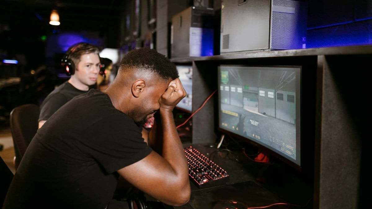 Two adult men experiencing disappointment while playing PC games in a modern indoor gaming arcade setting.