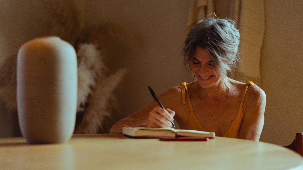 A senior woman sitting at a table, journaling happily in her cozy home office.