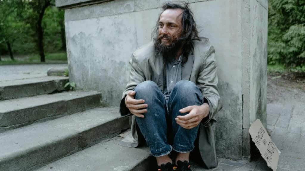 A homeless man sitting on concrete steps looking contemplative in an urban park setting.