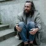 A homeless man sitting on concrete steps looking contemplative in an urban park setting.