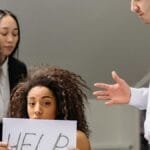 Woman holding HELP sign surrounded by colleagues in a tense office situation. via Pexels