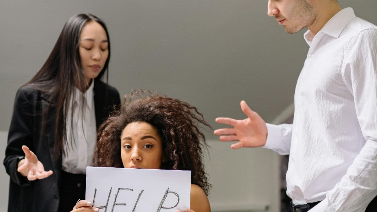 Woman holding HELP sign surrounded by colleagues in a tense office situation. via Pexels