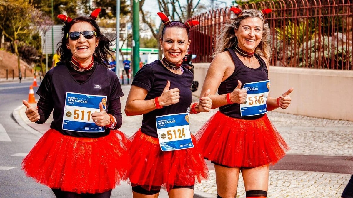 Three women in red tutus and devil horns joyfully run a marathon, smiling and giving thumbs up.