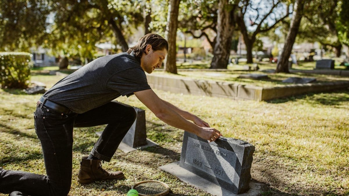 A man kneels at a cemetery tombstone with a tennis racket and ball nearby, expressing remembrance.