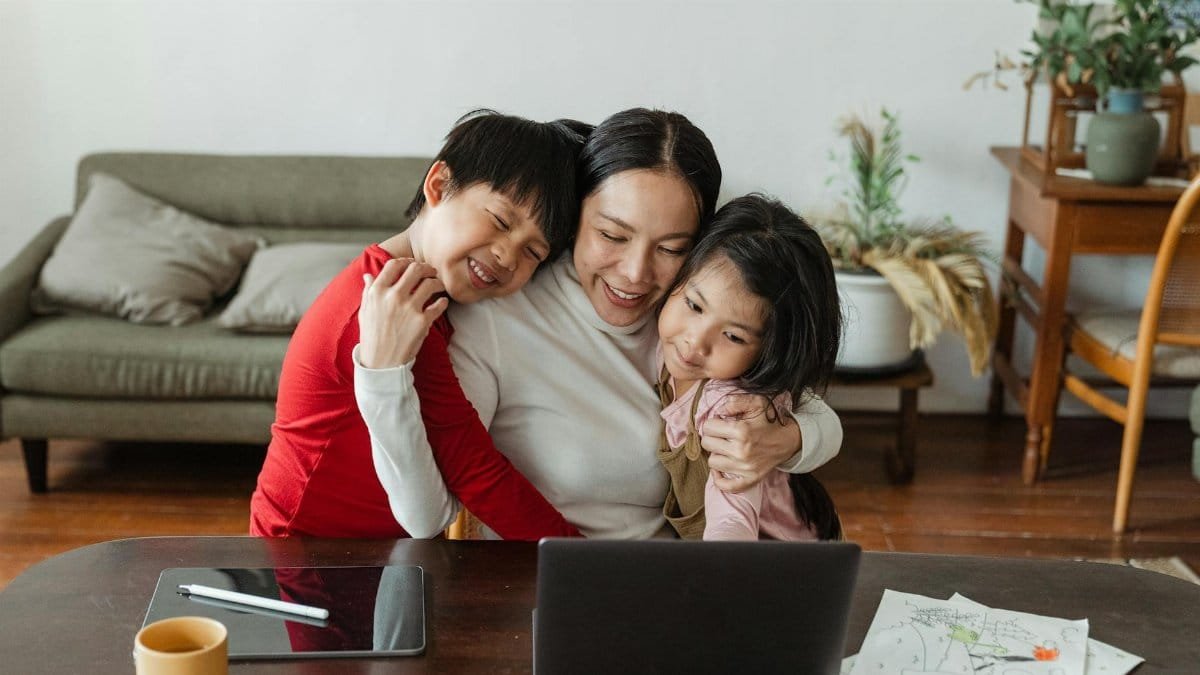 A mother embraces her children while working from home, blending family time with remote work.