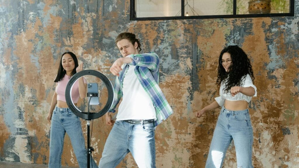 Group of friends dancing indoors with a smartphone and ring light setup for social media content. via Pexels