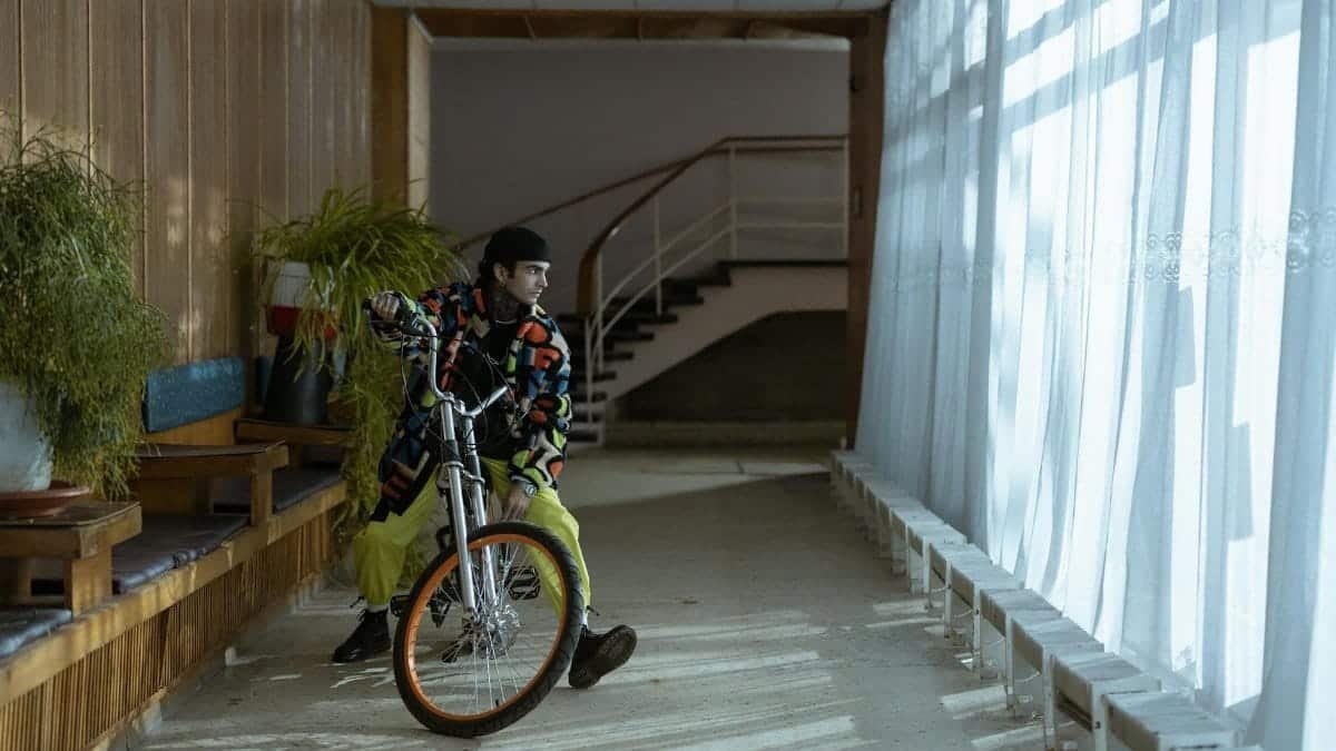 A fashionable man in vibrant clothing leans casually on a bike in a sunlit hallway.