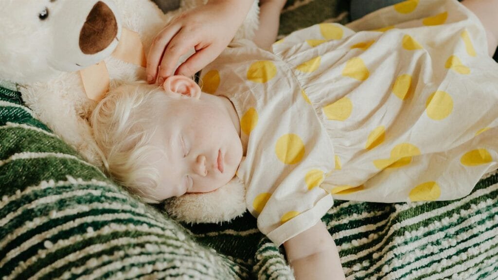 Adorable toddler with yellow polka dot dress sleeping on a sofa with a teddy bear.