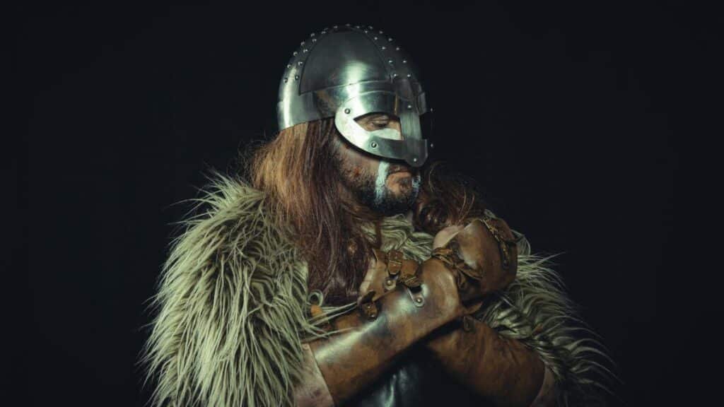 Portrait of a Viking warrior wearing a metal helmet and fur cloak, captured against a dark background.