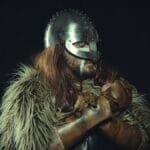 Portrait of a Viking warrior wearing a metal helmet and fur cloak, captured against a dark background.