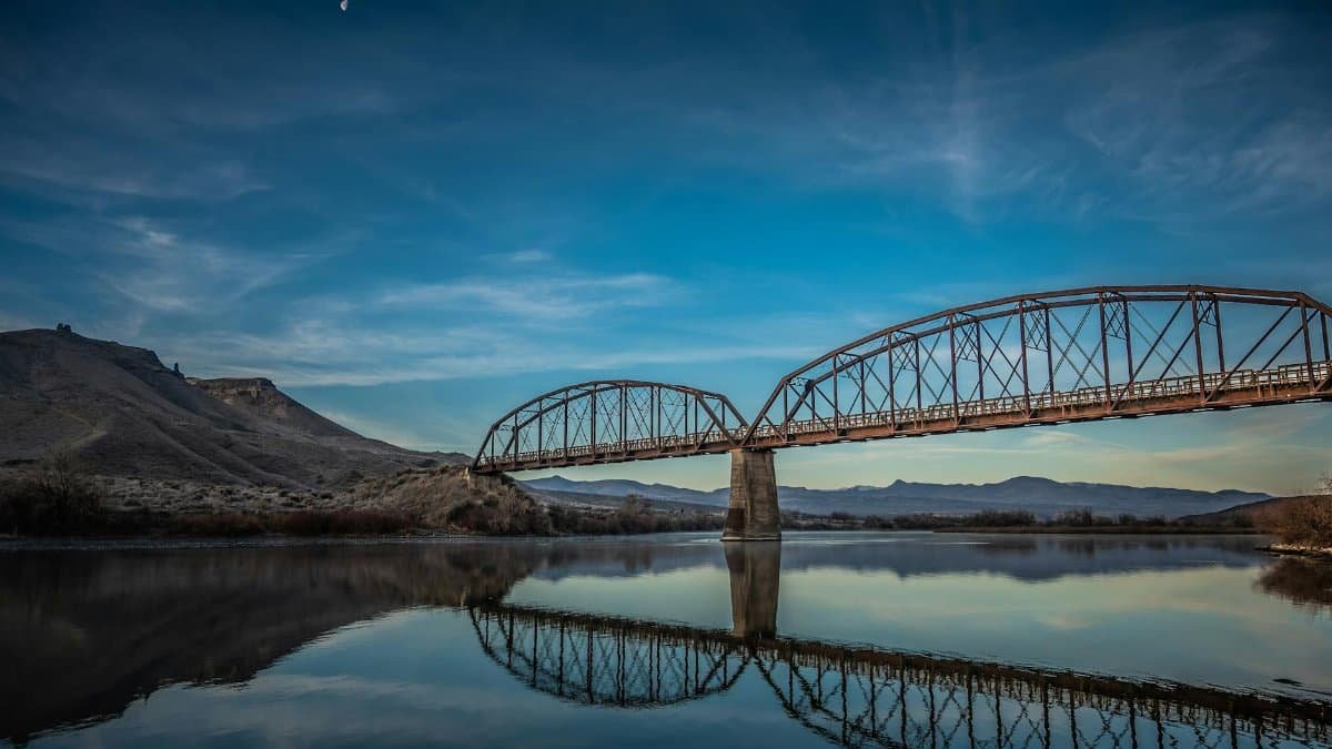 Scenic reflection of a steel bridge over a river at dusk with a moonlit sky.