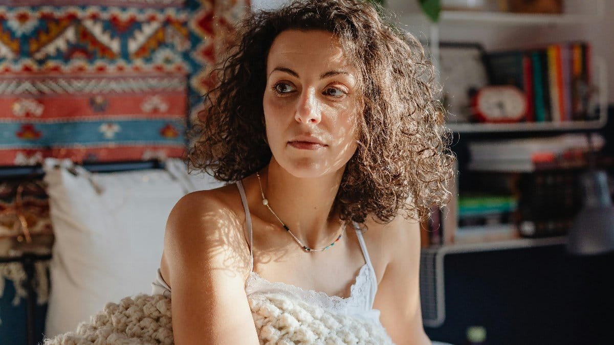 Woman sitting on bed wrapped in a knitted blanket, enjoying a cozy morning sun. via Pexels