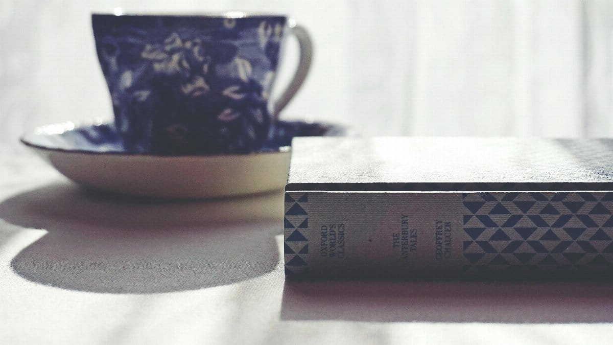 A vintage blue tea cup and a classic book on a white tablecloth, lit softly.
