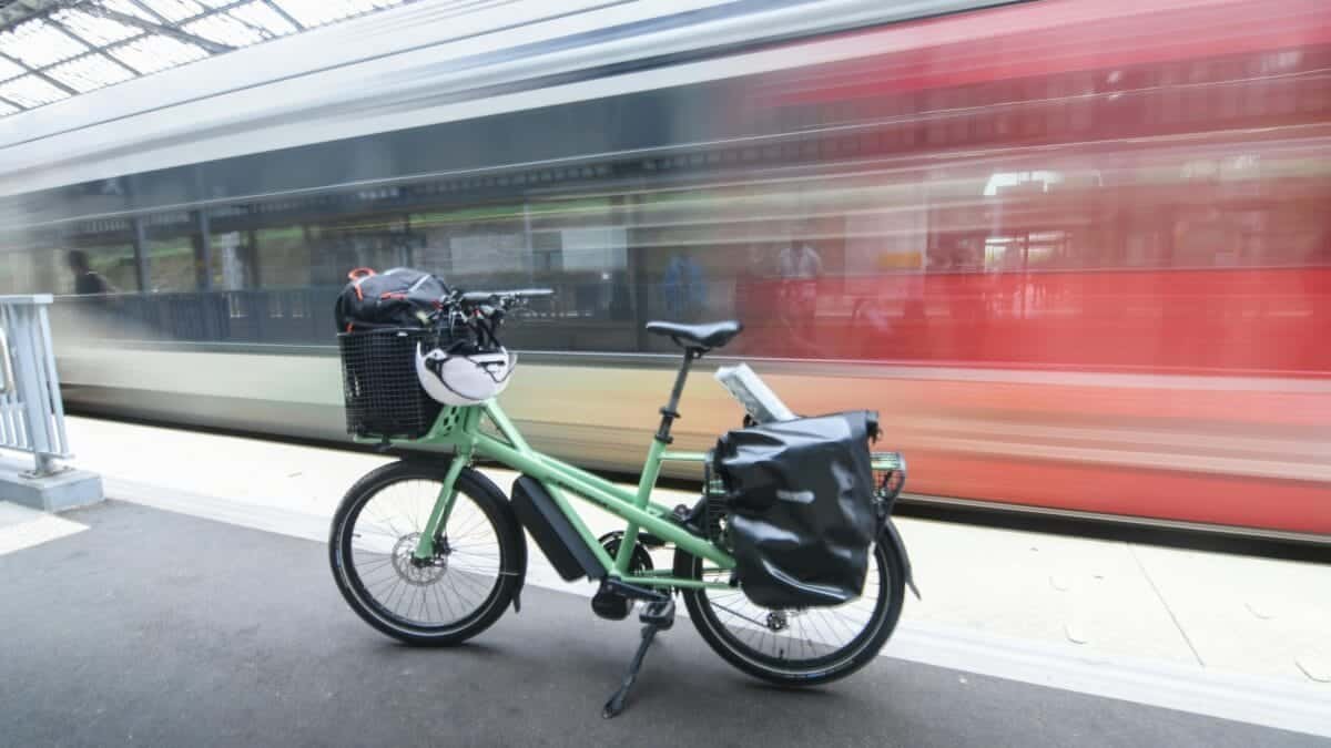 A green cargo bike parked at a train station platform in France, with a train motion blur in the background.
