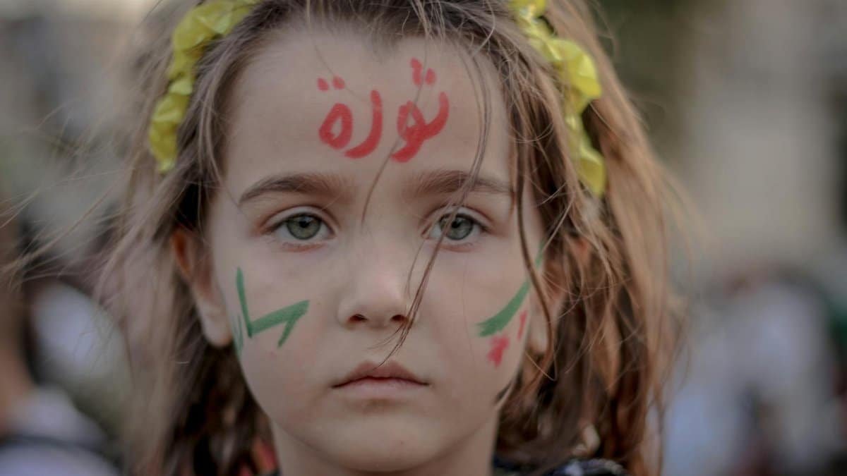 Unemotional little girl with symbols on cheeks and Arabic script on forehead looking at camera on blurred background