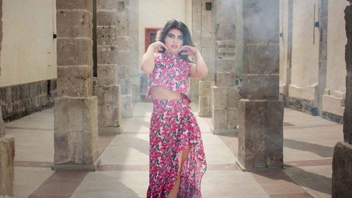 Model in vibrant floral dress poses amid ancient stone columns indoors.