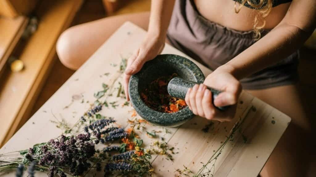 Close-up of hands using mortar and pestle to grind herbs, surrounded by dried flowers on wooden surface.