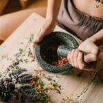 Close-up of hands using mortar and pestle to grind herbs, surrounded by dried flowers on wooden surface.