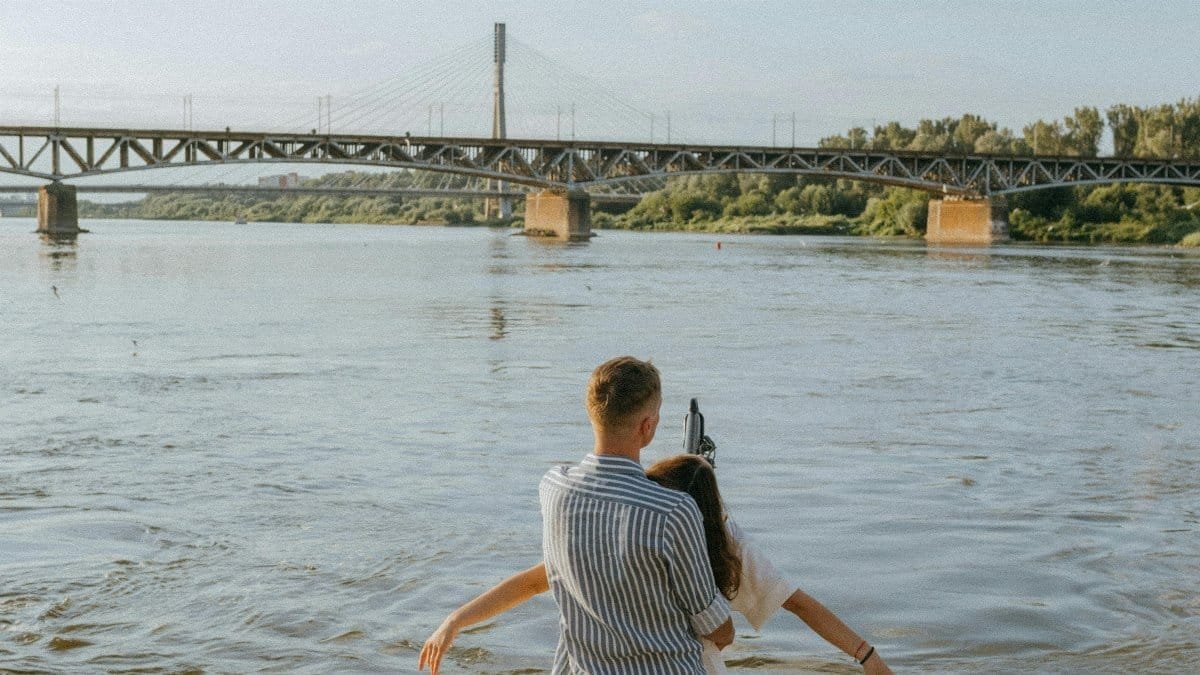 Couple embracing on a boat, sailing under a bridge over a river during a sunny day.