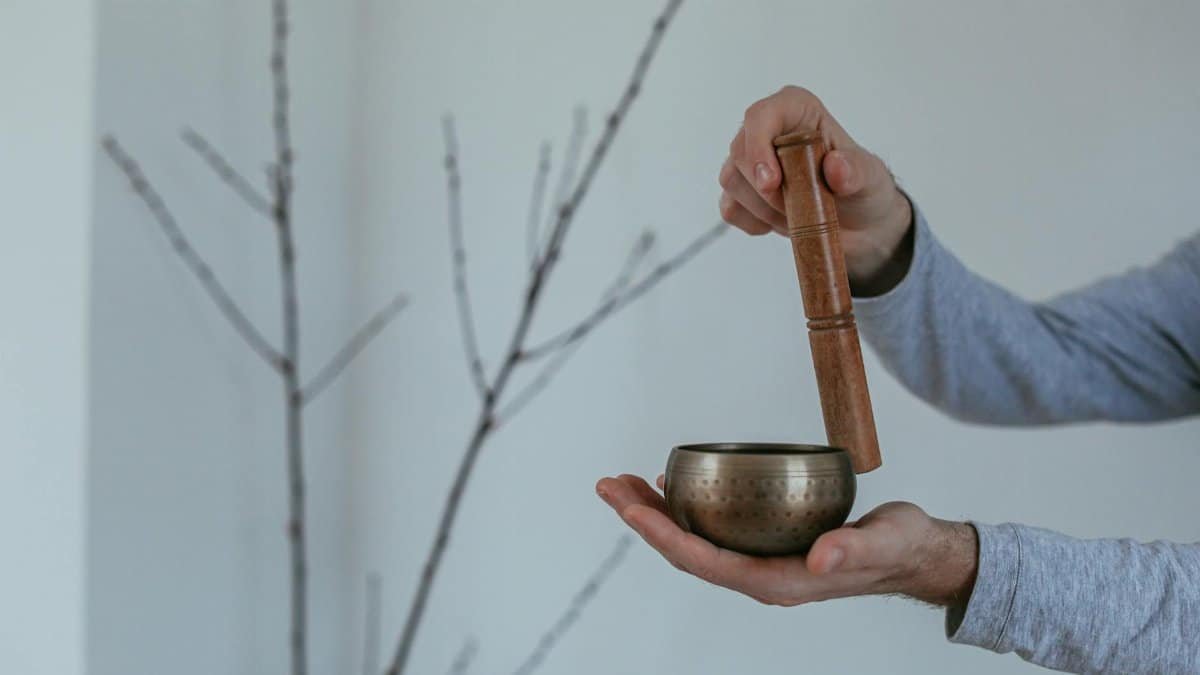 Hands holding a brass singing bowl with a wooden mallet against a minimalist indoor background.