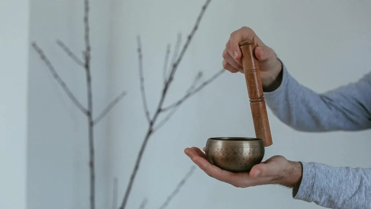 Hands holding a brass singing bowl with a wooden mallet against a minimalist indoor background.