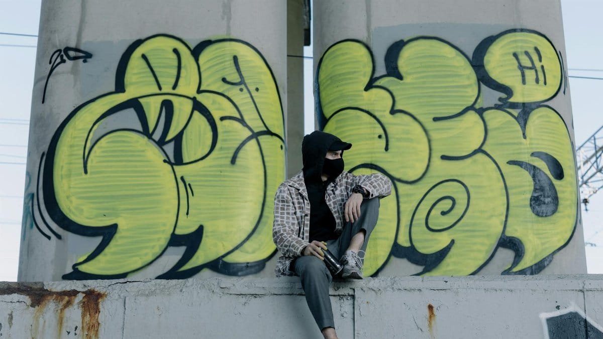 A street artist sits by vibrant graffiti under an urban bridge.