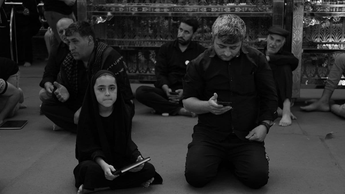 A group of individuals sitting indoors, some praying while others use digital devices, in a calm and contemplative setting.