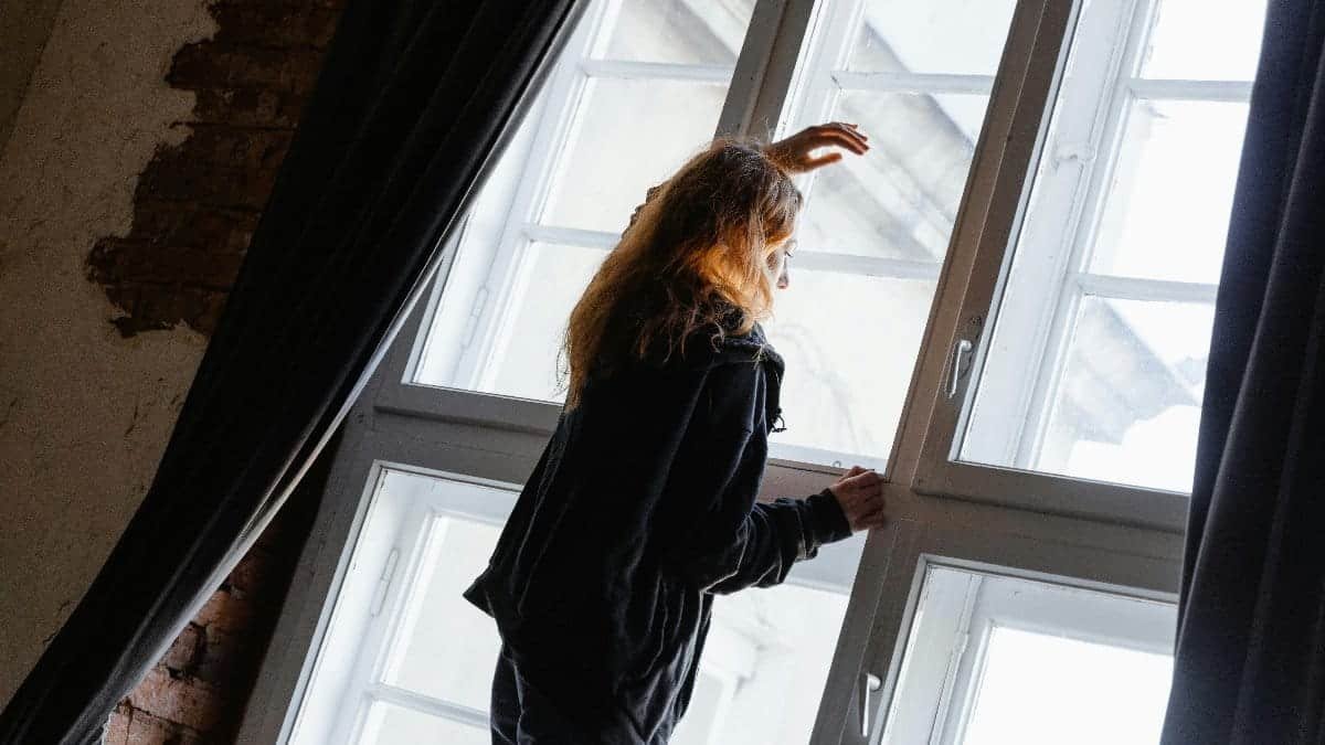 A woman stands in deep thought near a large window, embracing solitude.