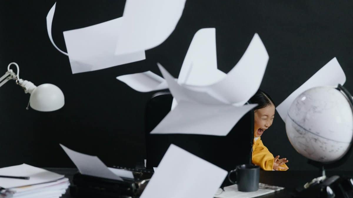 A child laughs as papers fly around a dark office setting, creating a sense of chaos.