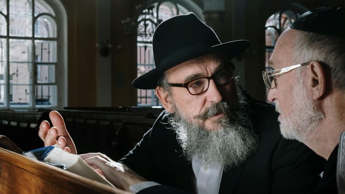 Two senior Orthodox men engaged in a deep conversation over a religious text indoors.