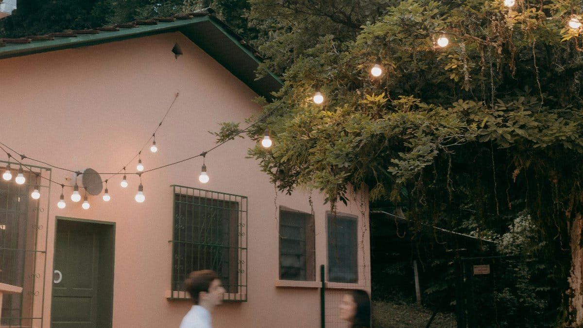 A couple walks past a lit tree and house in a tranquil courtyard, evoking a serene evening atmosphere. via Pexels