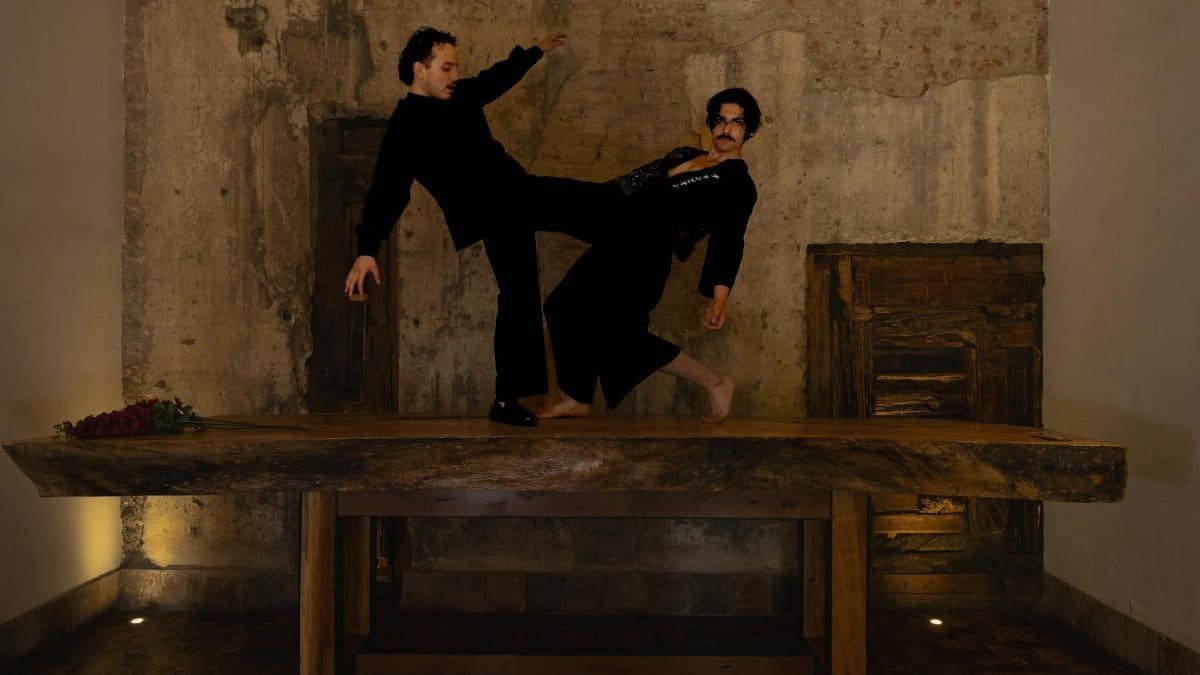 Two men perform an expressive movement atop a rustic wooden table indoors, capturing a moment of balance and art.