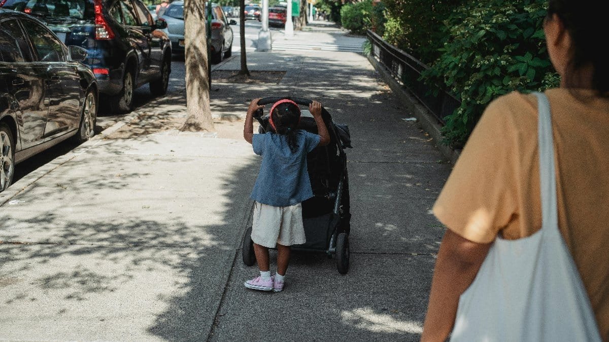 A young girl joyfully pushes a stroller on a sunlit city sidewalk with mom nearby.