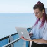 A young woman works remotely on her laptop by the sea on a sunny day, embodying the digital nomad lifestyle. via Pexels