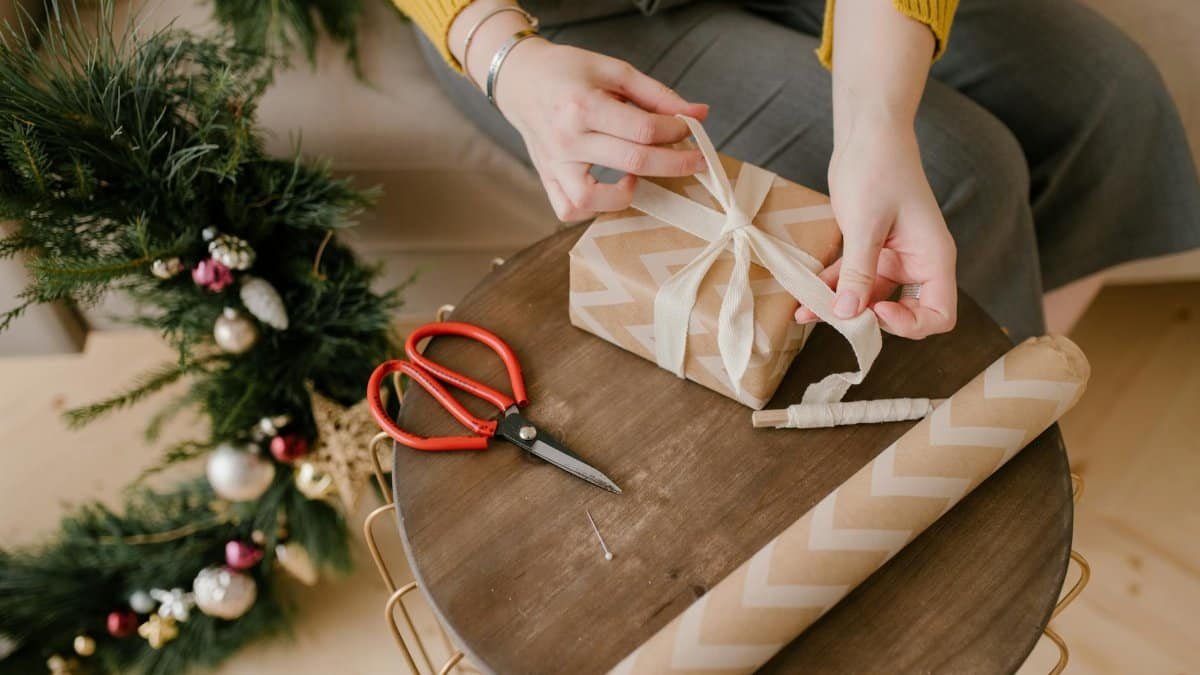 Close-up of hands tying a bow on a gift box with holiday decorations around.