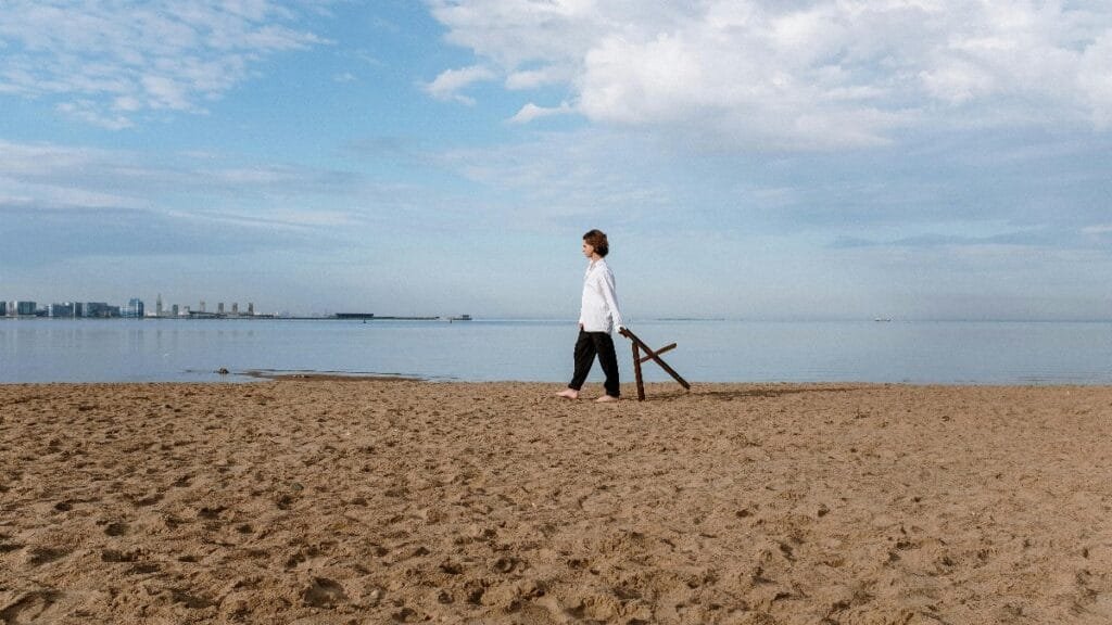 Man in white shirt walking on sandy beach under blue sky, serene seascape. via Pexels