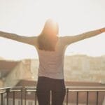 A woman stands with outstretched arms on a sunny balcony, embracing the morning light.