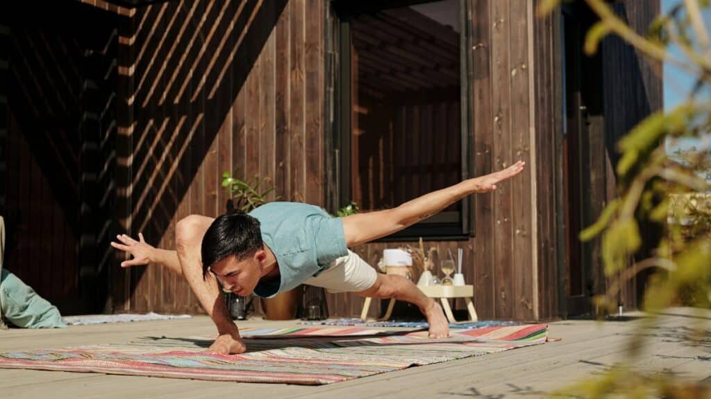 A man performs an advanced yoga pose on a colorful mat outside on a sunny veranda. via Pexels